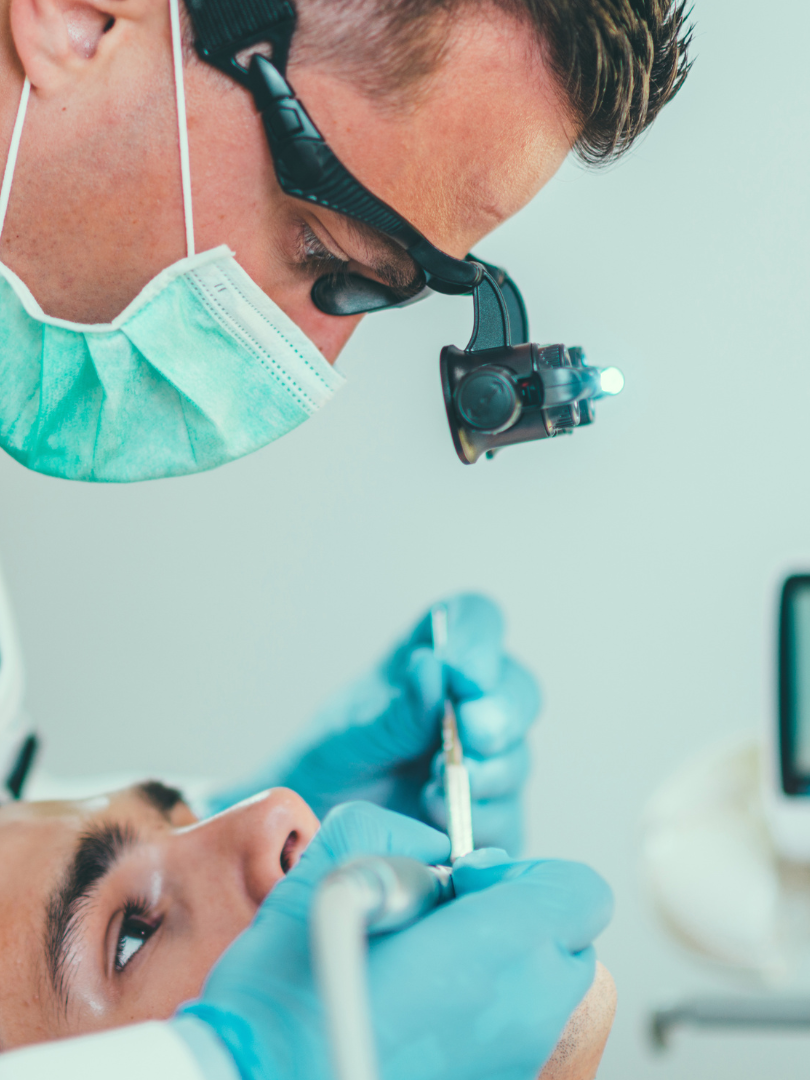 Dentist in mask, glasses, and gloves, examining a patient's teeth with tools in a clinical setting.