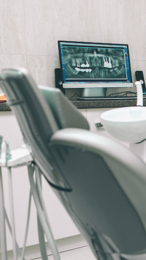 Dental chair in a bright room with a digital x-ray of teeth on a monitor in the background.