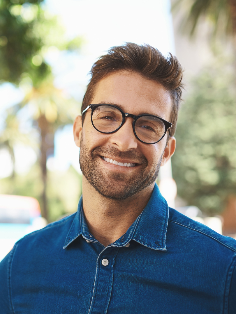 Smiling man with glasses and a beard, wearing a blue shirt, outdoors.