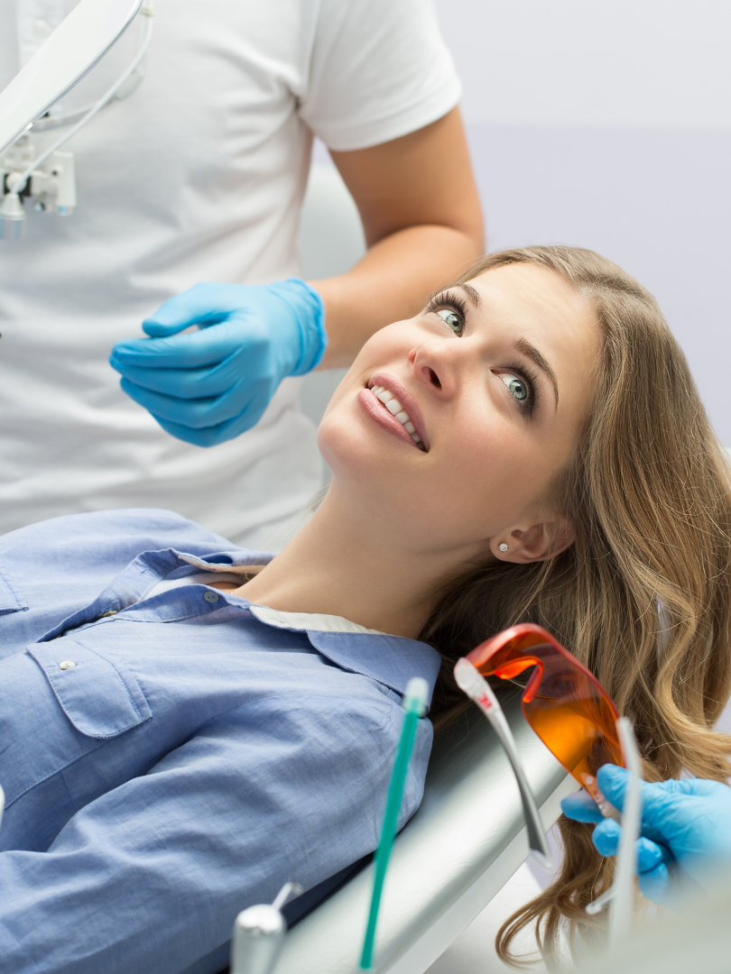 Woman at dentist, smiling, looking up, blue-gloved hands, orange protective glasses, dental setting.