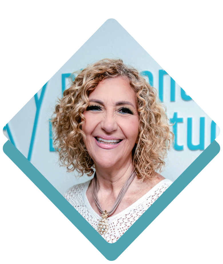 Woman with curly blonde hair smiles at the camera, wearing a white shirt and necklace.