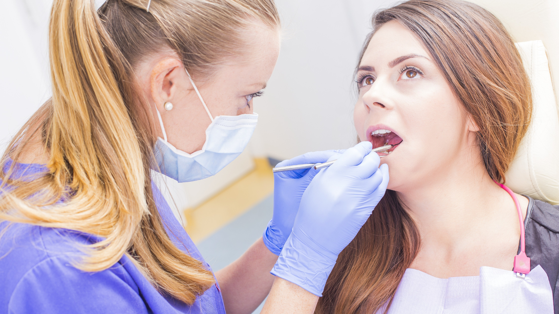 A dentist wearing a blue uniform, mask, and gloves examines a patient's teeth in a clinical setting.