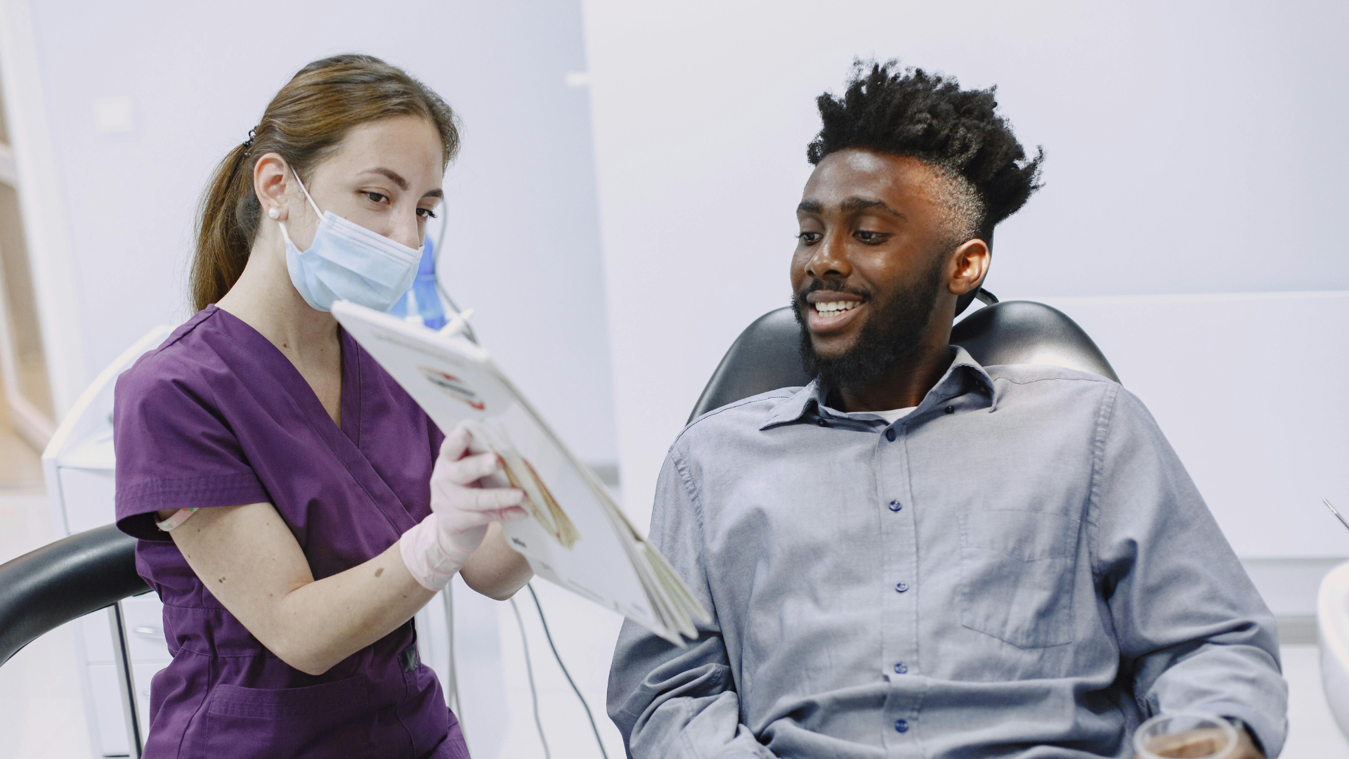 Dentist in purple scrubs shows a tablet to a smiling patient in a dental chair