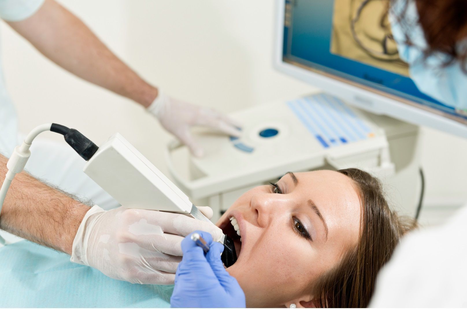 Dentist using scanner on patient's mouth, with a screen showing teeth scan; white gloves, blue gloves.