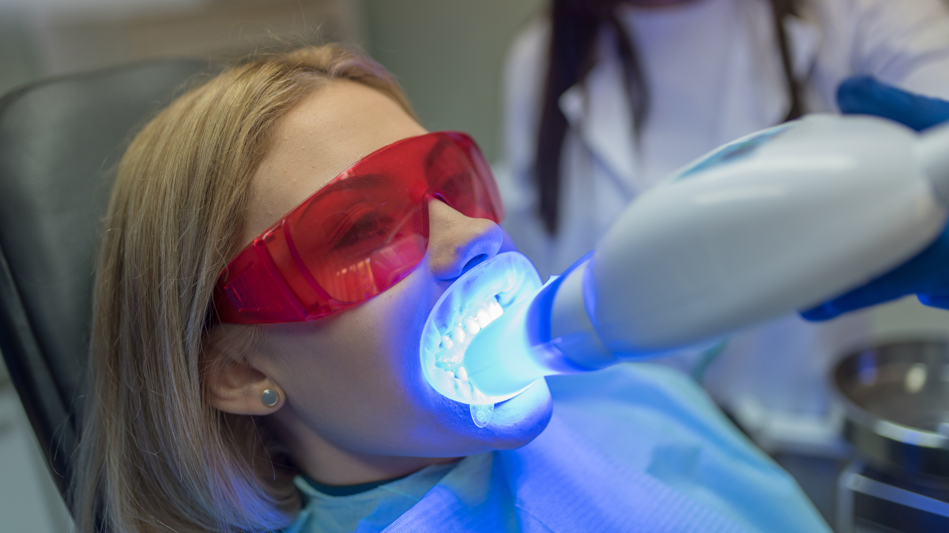 Woman undergoing teeth whitening with blue light in a dental office, wearing safety glasses.