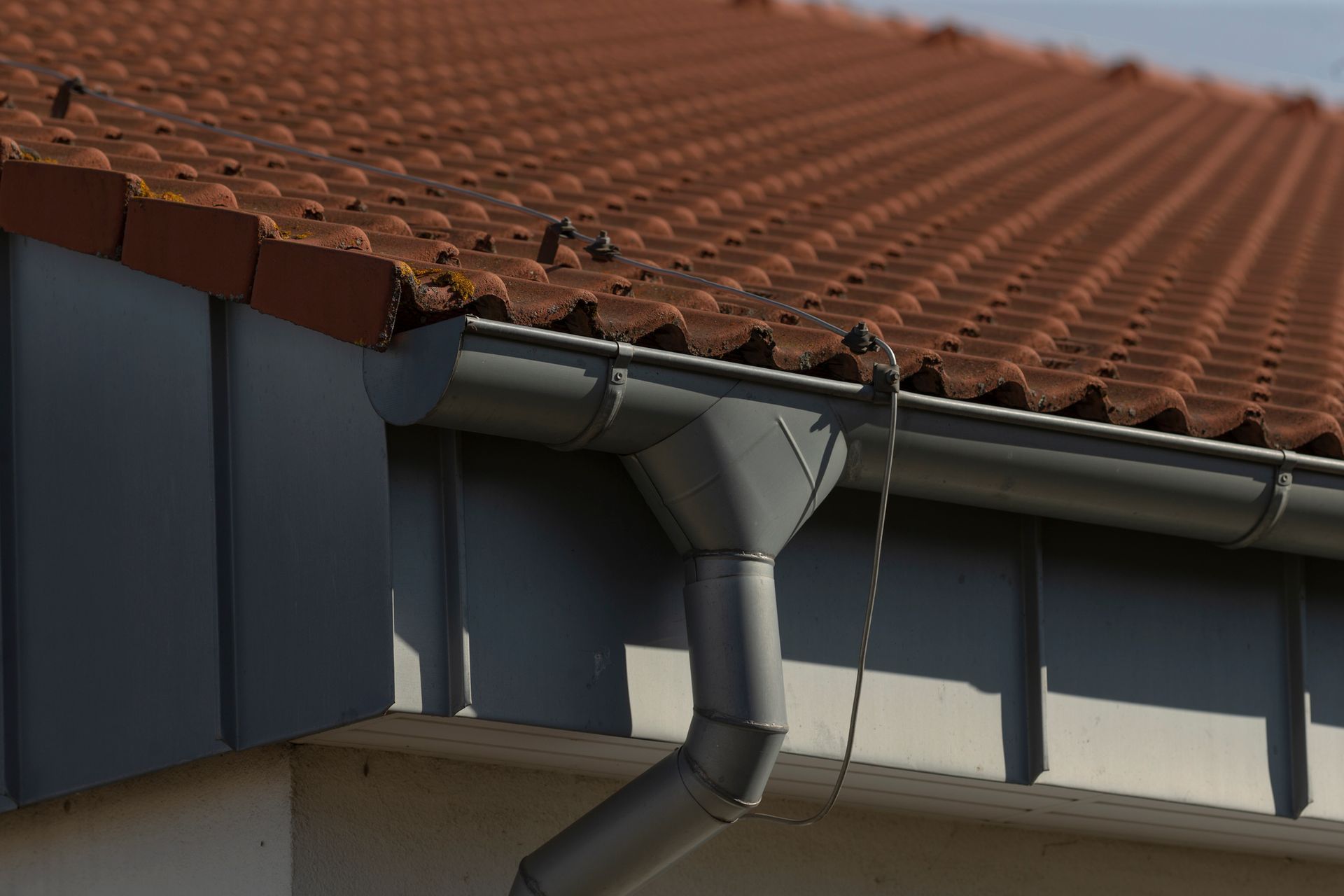 Gray gutter and downspout on a roof with red-brown tiles.