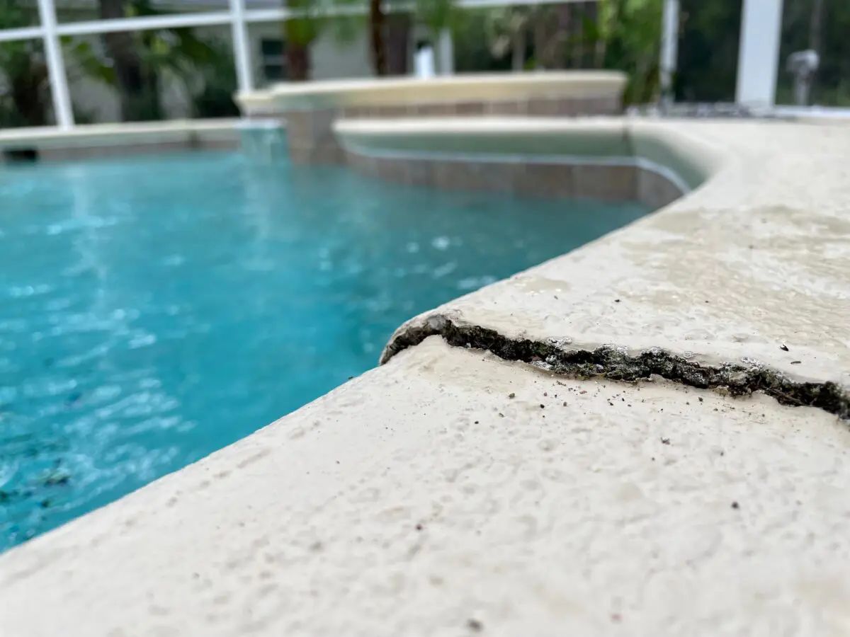 Close-up of a pool's edge with cracked concrete, turquoise water, and a blurred background of a house and greenery.