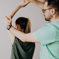 A man in glasses assisting a woman in a dark top with an overhead stretch in a studio
