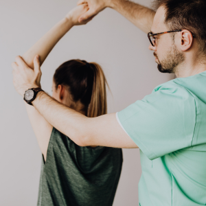 A man in glasses assisting a woman in a dark top with an overhead stretch in a studio