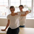 Therapist guiding a seated woman through a shoulder stretch in a bright studio