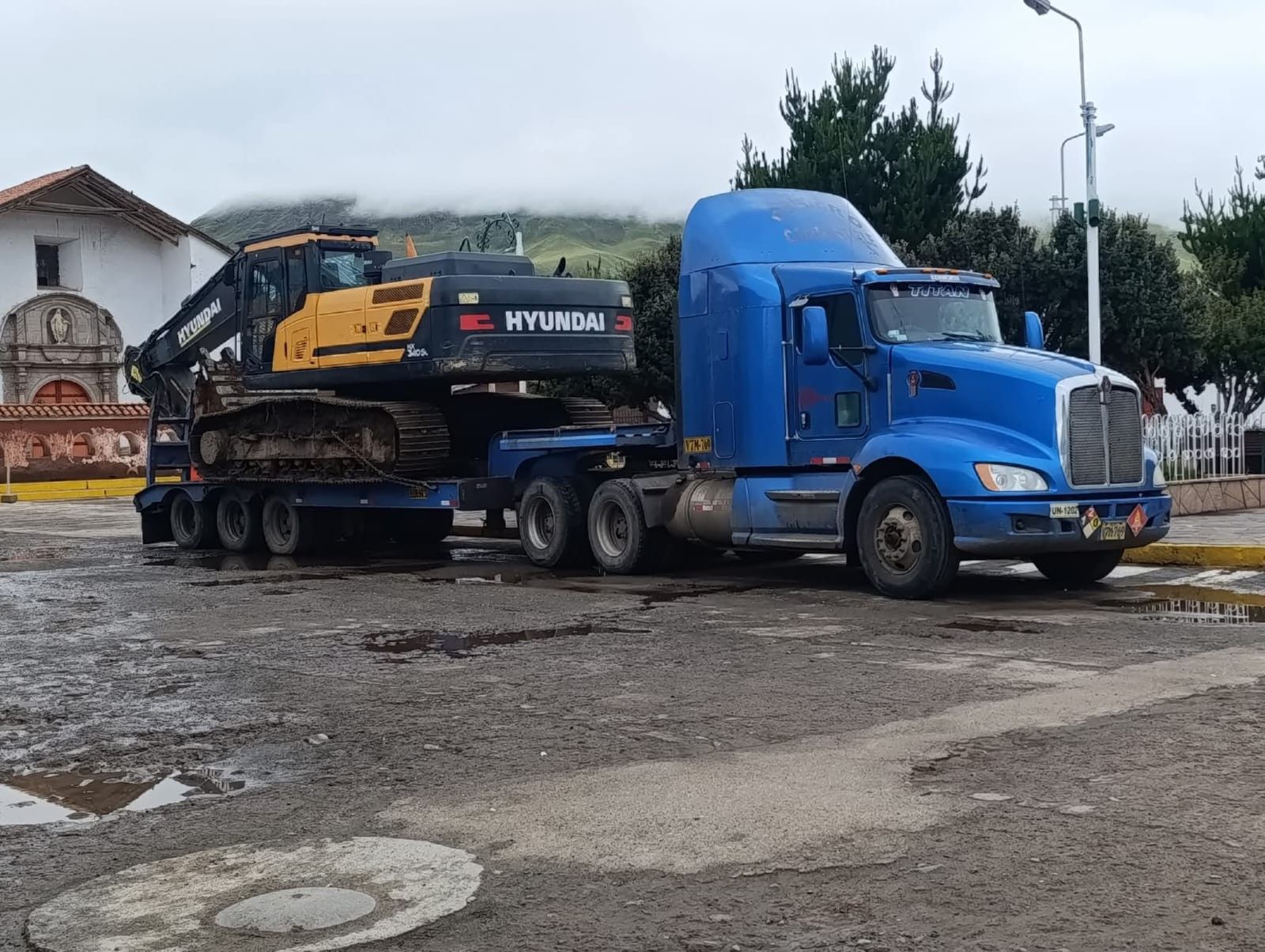 Un camión semirremolque azul transporta una excavadora amarilla en un remolque.