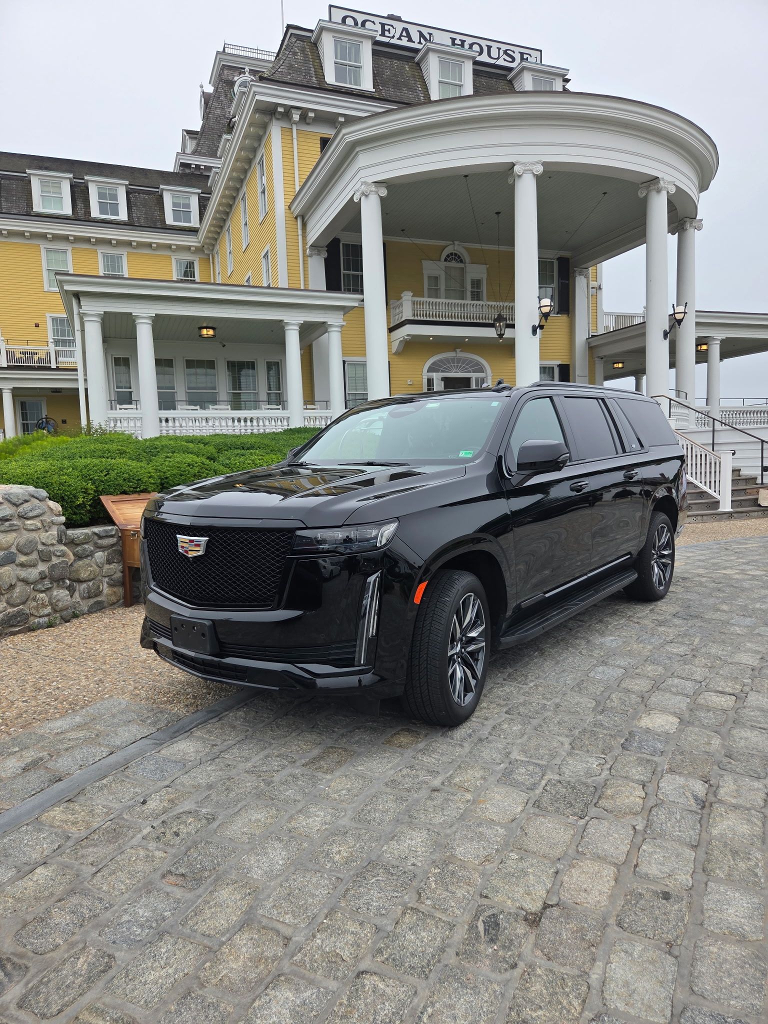 A black Cadillac Escalade parked on a stone driveway in front of a yellow historic hotel with white columns.