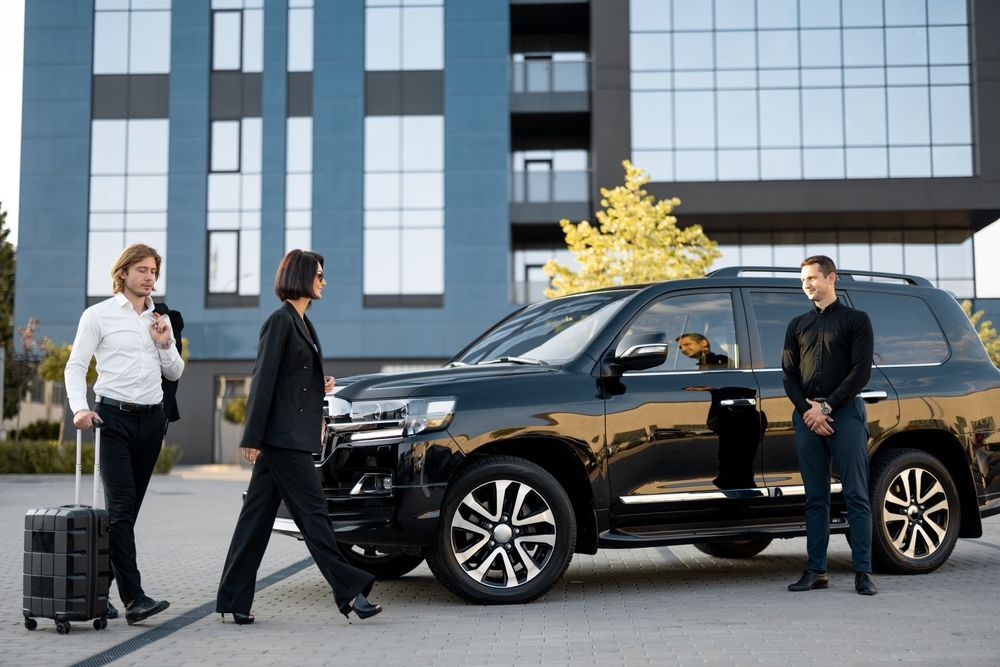 Three people near a black SUV parked outside a modern building, with one person holding a rolling suitcase.
