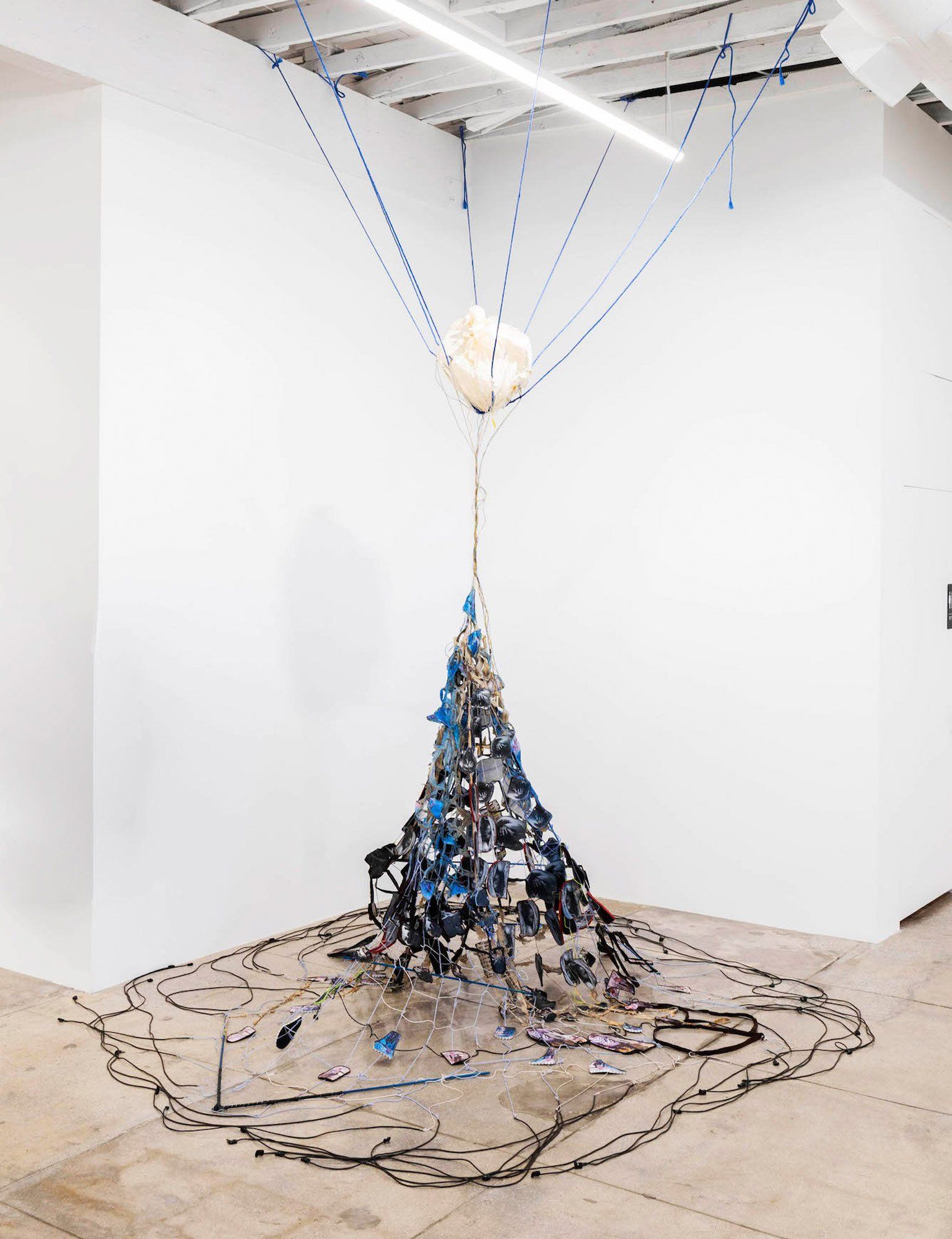 A sculpture of a christmas tree made out of trash is hanging from the ceiling in a room.