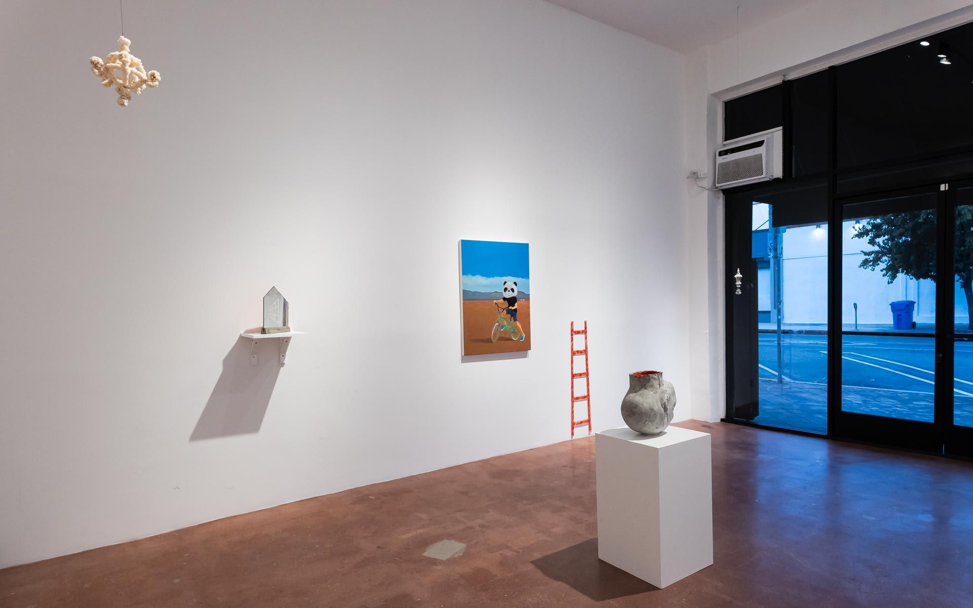 Painting of a figure with white flowers beside title wall text, a gray ceramic vessel on a pedestal, a black staircase and a blue house-shaped shelf work. Installation view, Beryl Odette: Seaing, Track 16.