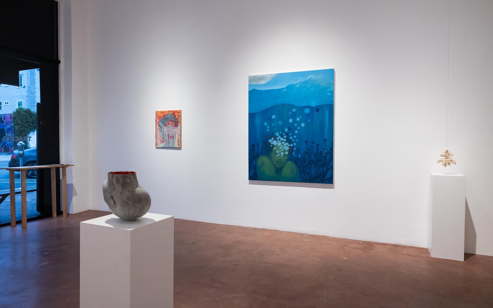 Blue painting of flowers under water at left, a small hanging sculpture on a pedestal, and a large portrait painting beside title wall text. Installation view, Beryl Odette: Seaing, Track 16