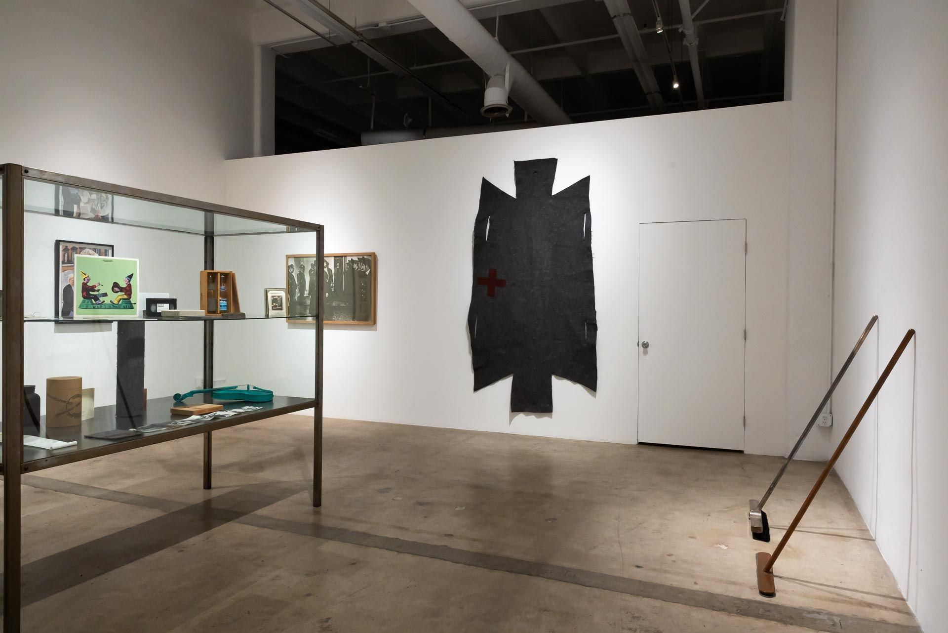 Museum gallery with framed artworks, a large dark wall piece, and two long-handled tools leaning on the right wall. TRACK 16 | DOWNTOWN LA art exhibition and retrospective of JOSEPH BEUYS titled POTENTIAL GOODS. Sculpture, mixed media and drawings.