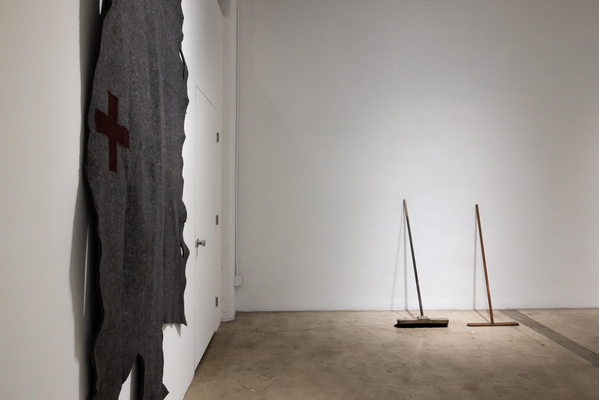 Damaged wall with a red cross and two slender sculpture stands in a bare room. TRACK 16 | DOWNTOWN LA art exhibition and retrospective of JOSEPH BEUYS titled POTENTIAL GOODS. Sculpture, mixed media and drawings.