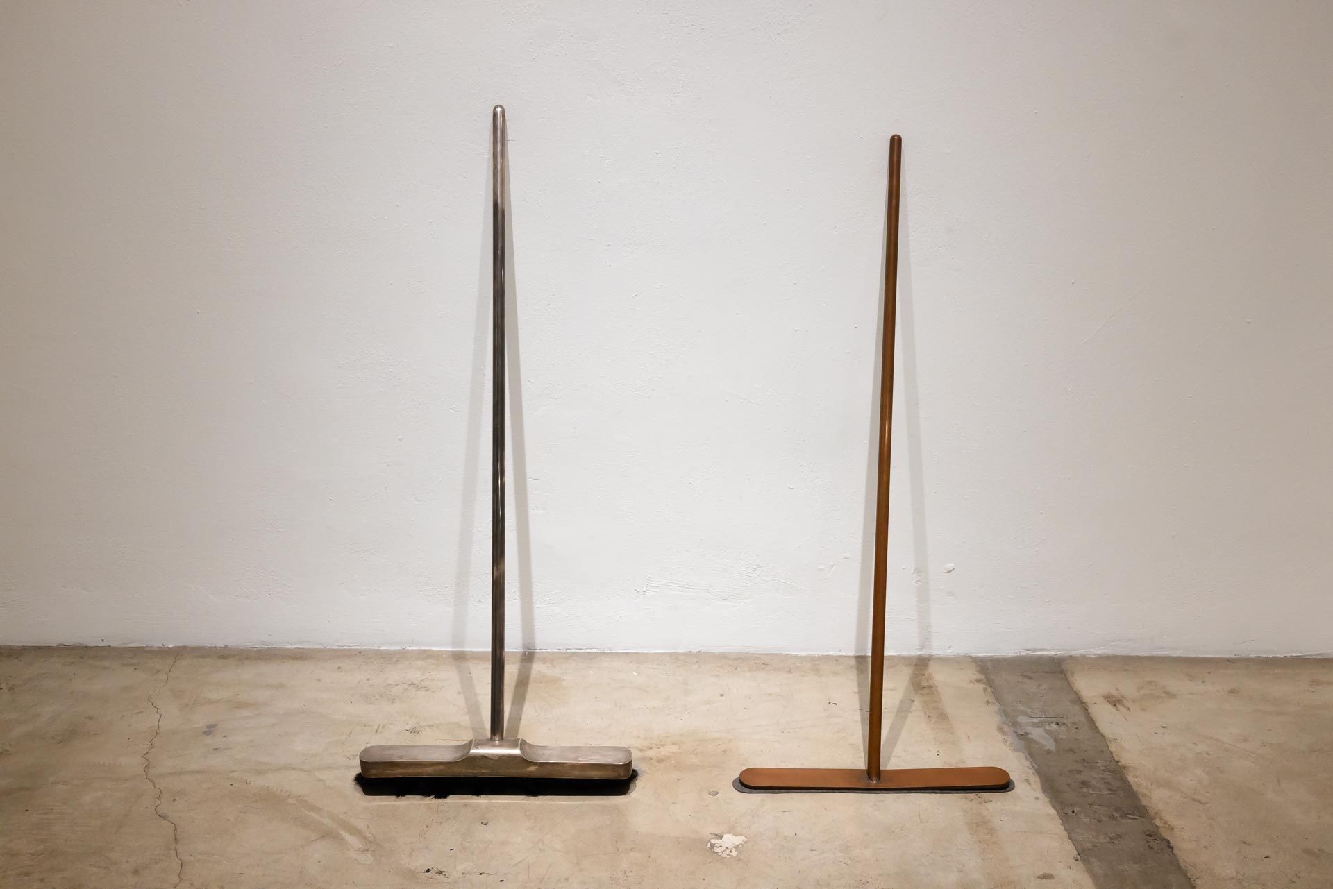 Two brooms standing upright against a plain wall, one dark brown and one light brown. TRACK 16 | DOWNTOWN LA art exhibition and retrospective of JOSEPH BEUYS titled POTENTIAL GOODS. Sculpture, mixed media and drawings.