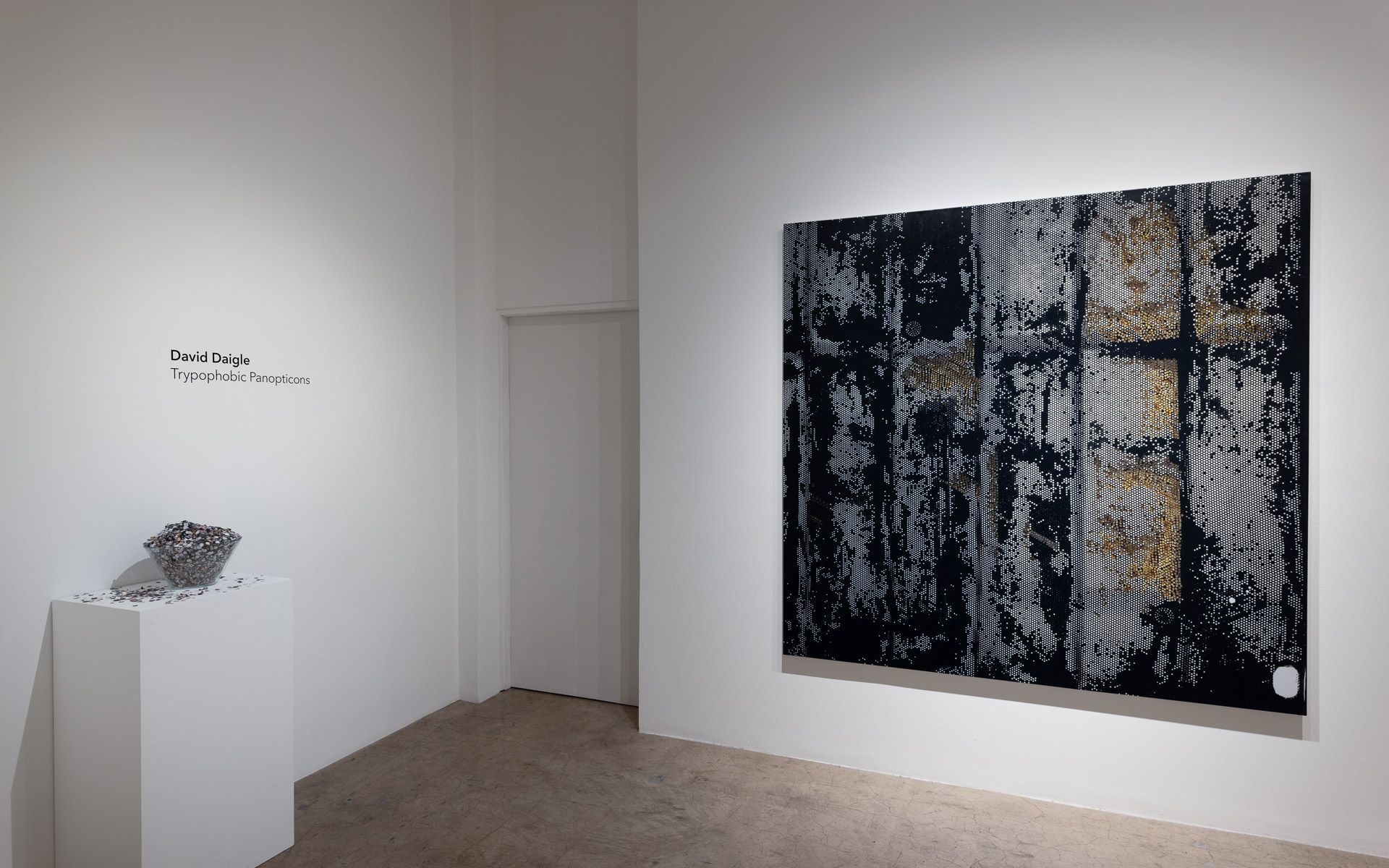 Large dark perforated panel with silver and gold marks on a white wall, a small metal sculpture on a white plinth and title wall text at left. Installation view, David Daigle: Trypophobic Panopticons, Track 16.