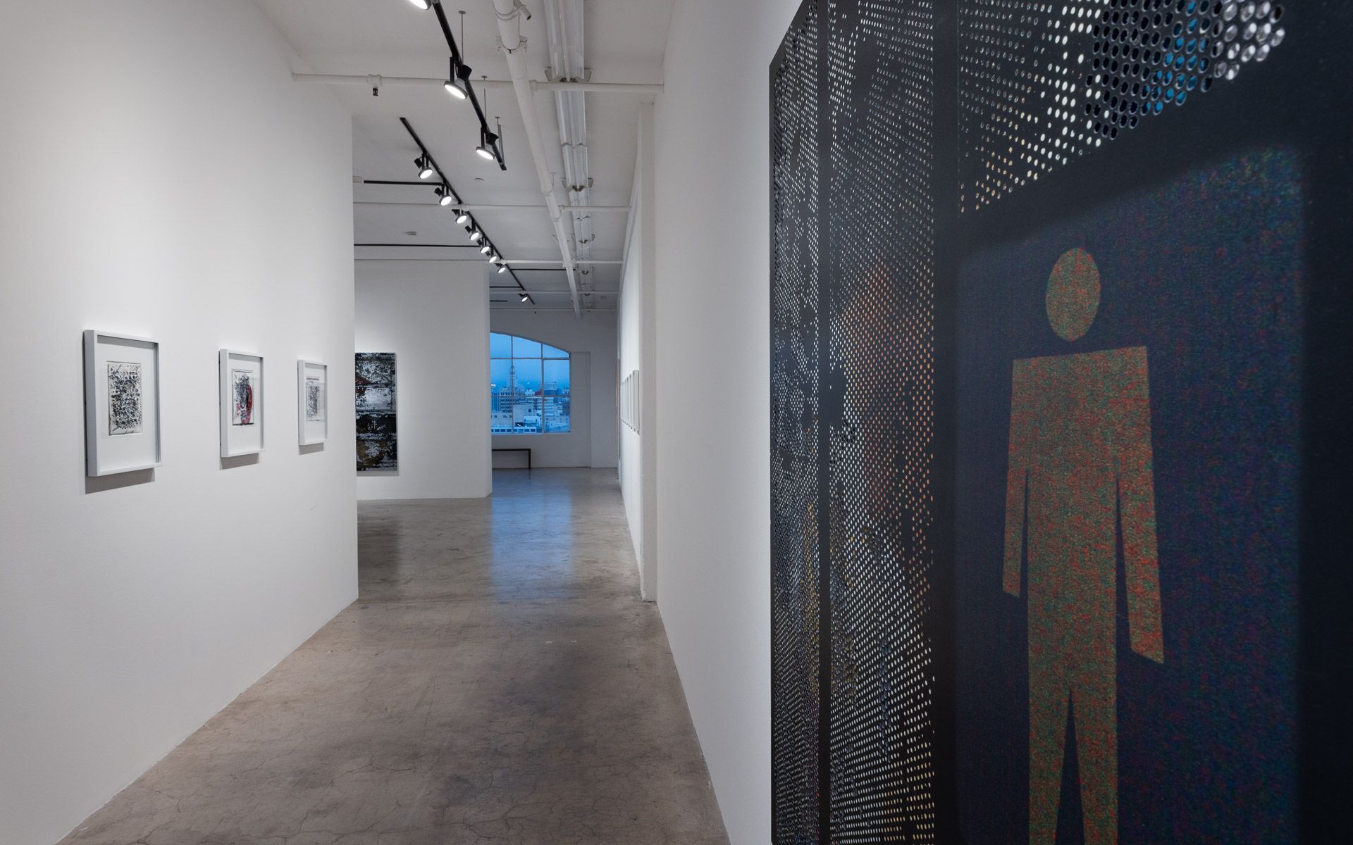 Close view of a dark perforated panel with a gold standing-figure pictogram, with small framed works down the gallery beyond. Installation view, David Daigle: Trypophobic Panopticons, Track 16.