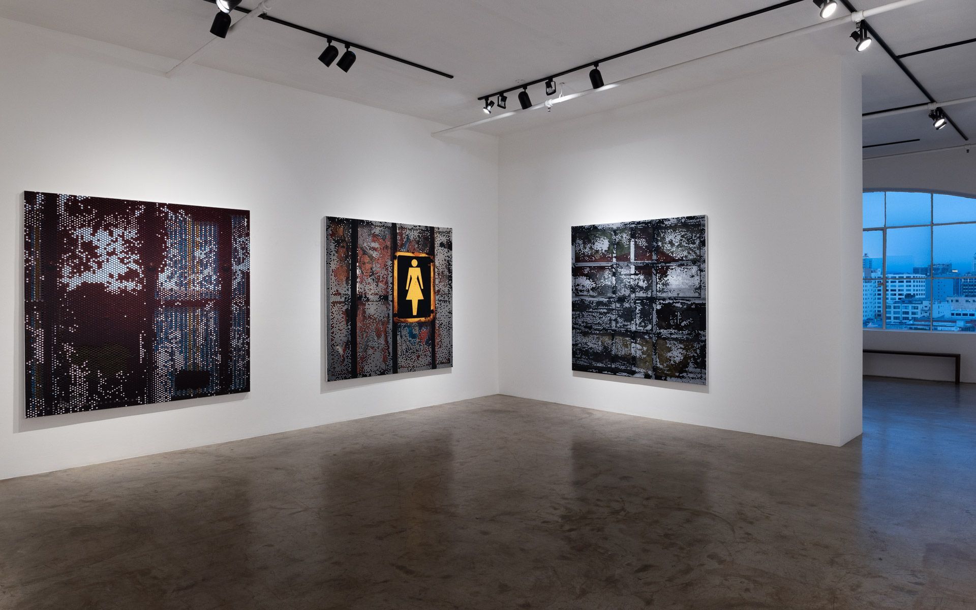 Three large dark perforated panels on white walls in a corner, one with a yellow woman pictogram sign, before a window at right. Installation view, David Daigle: Trypophobic Panopticons, Track 16.