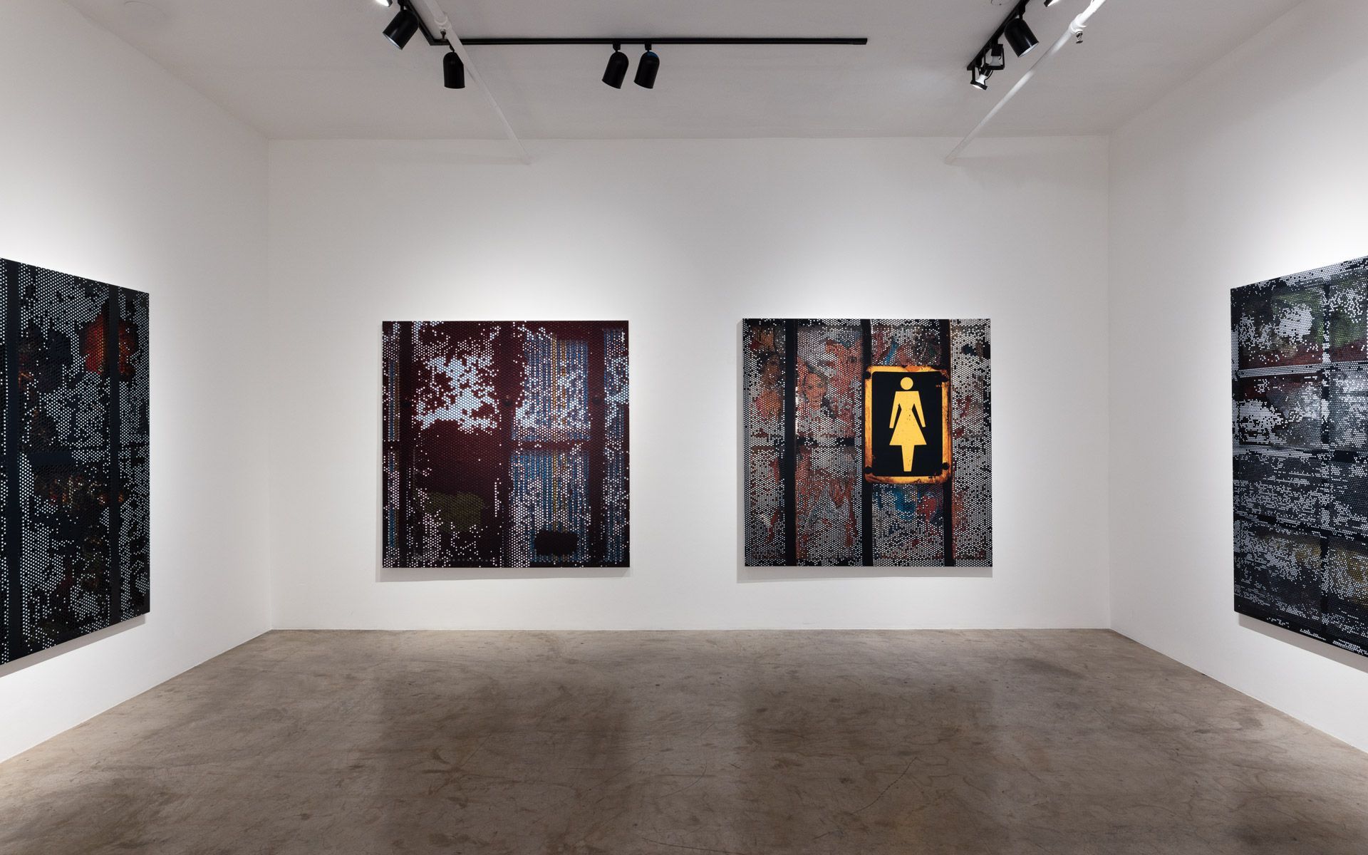 Three dark perforated panels across a white wall, the center one with a yellow woman pictogram, and another panel angled at the right edge. Installation view, David Daigle: Trypophobic Panopticons, Track 16.
