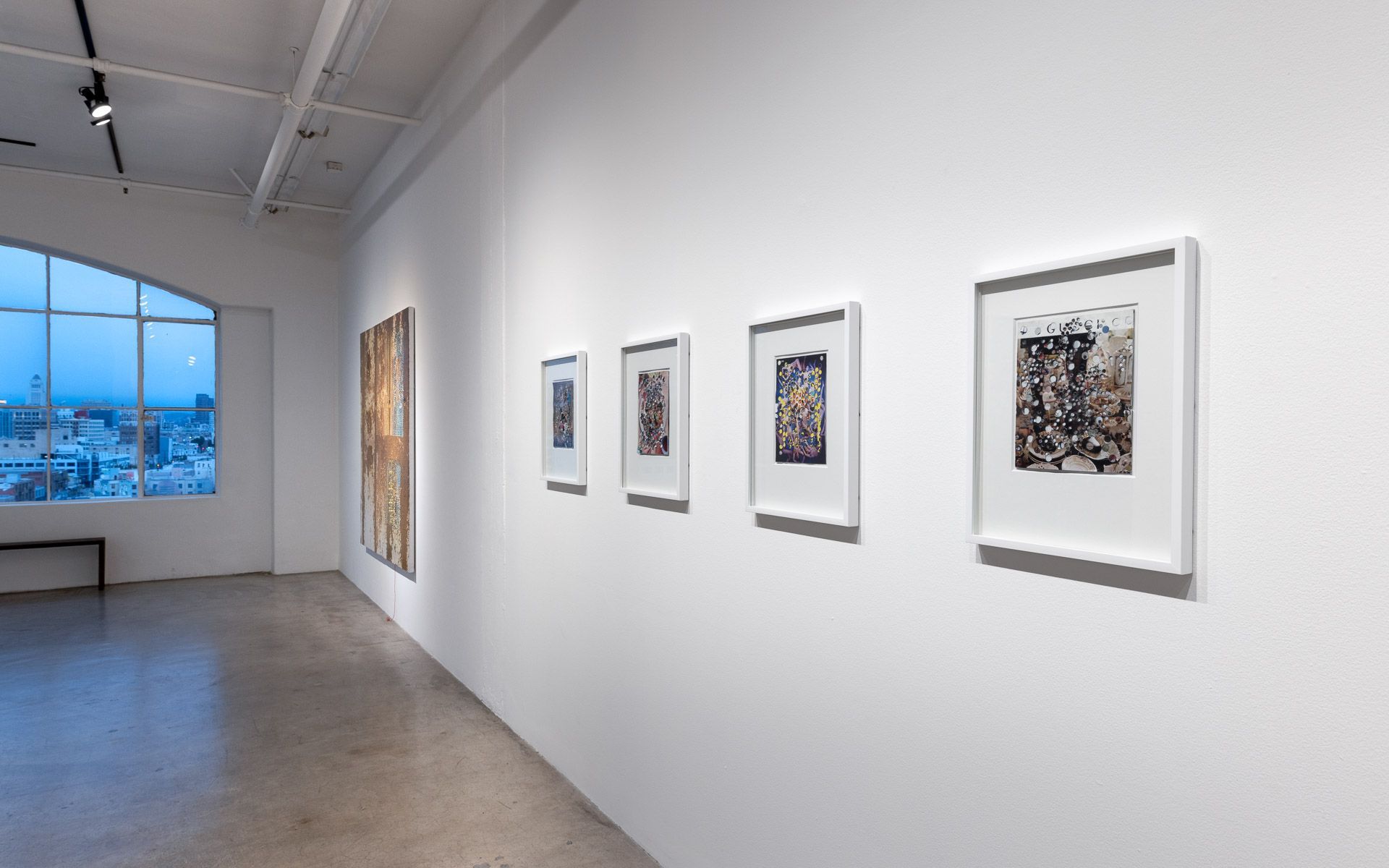 Row of small white-framed works on a long wall, a rusty-orange panel at left and an arched window at dusk. Installation view, David Daigle: Trypophobic Panopticons, Track 16.