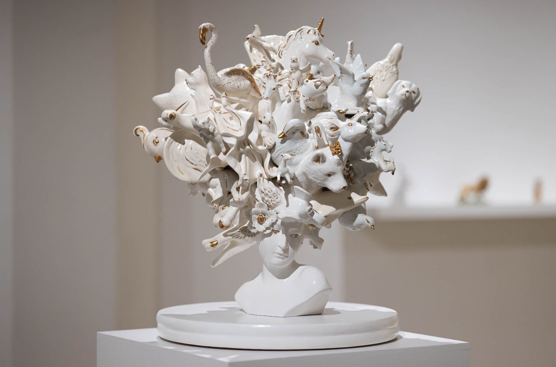 White sculpture of a bust crowned with a mass of white animals and flowers, on a round white base and pedestal. Installation view, Debra Broz: Strange World, Track 16.