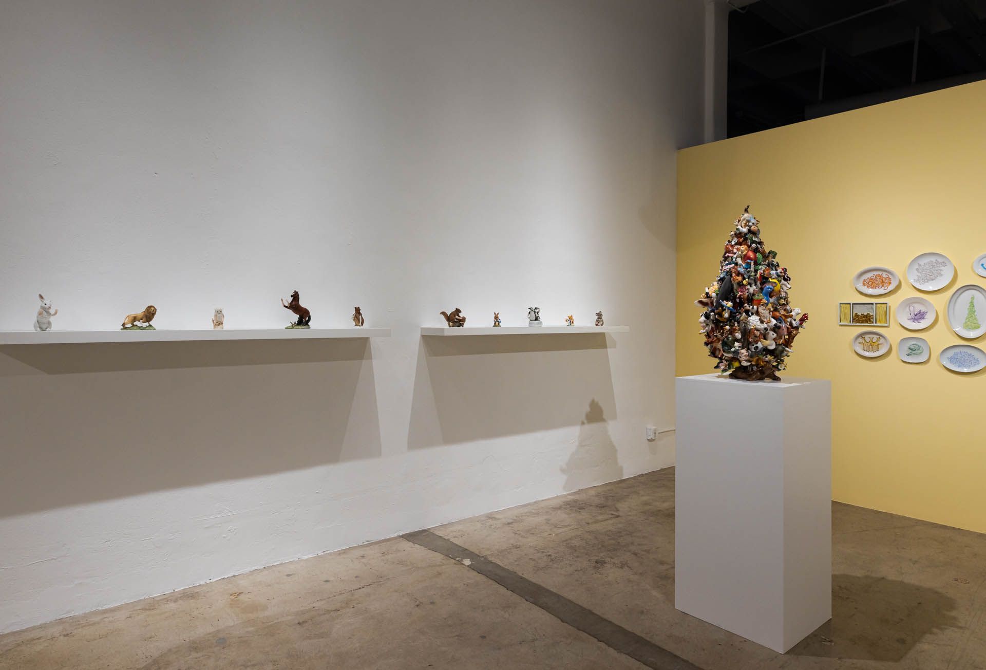 Long white wall with two shelves of small ceramic animals, a ceramic tree of colorful animals on a pedestal and a yellow wall with plates. Installation view, Debra Broz: Strange World, Track 16.