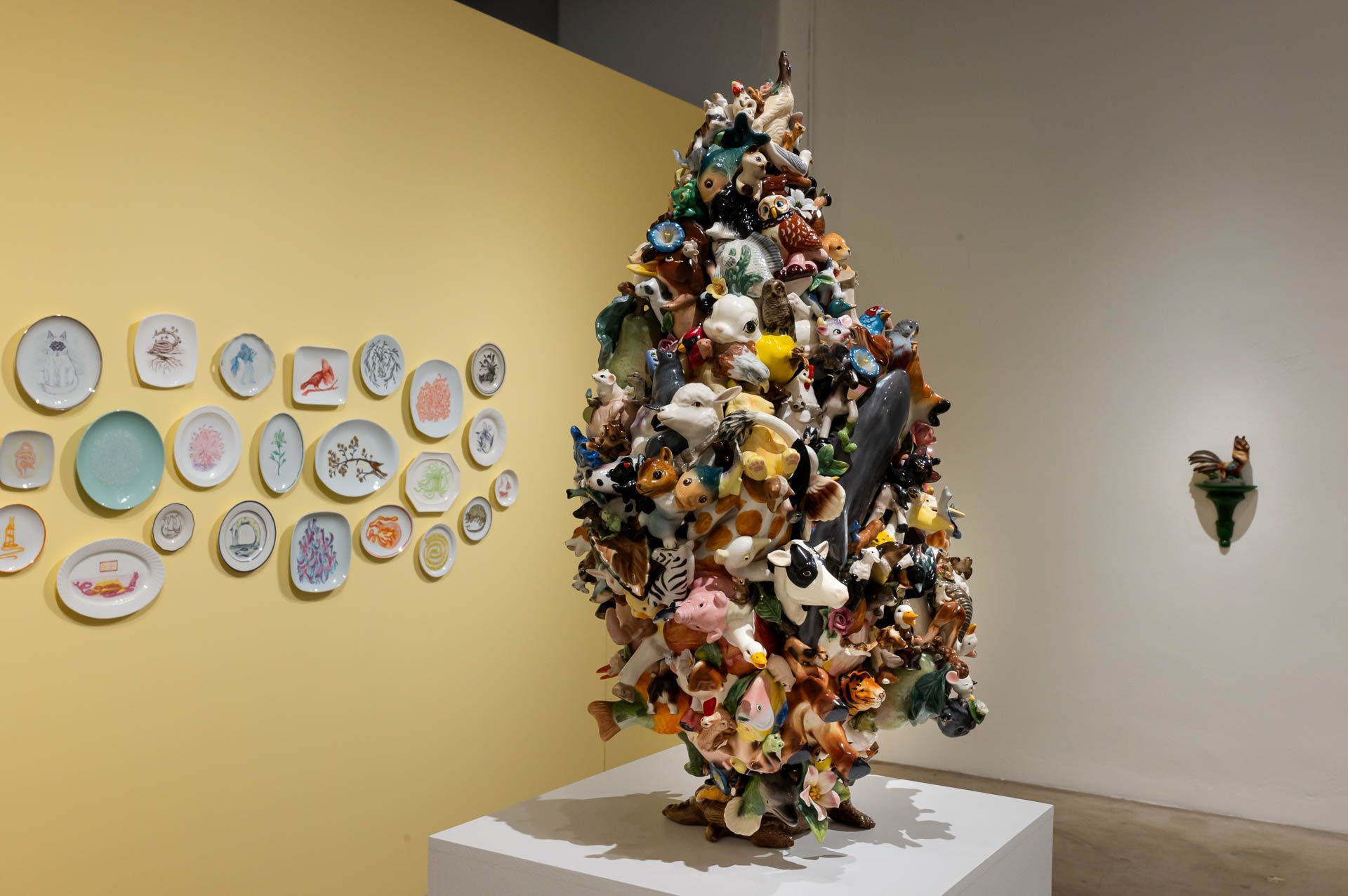 Tall ceramic sculpture of colorful animals on a white pedestal beside a yellow wall hung with painted plates. Installation view, Debra Broz: Strange World, Track 16.