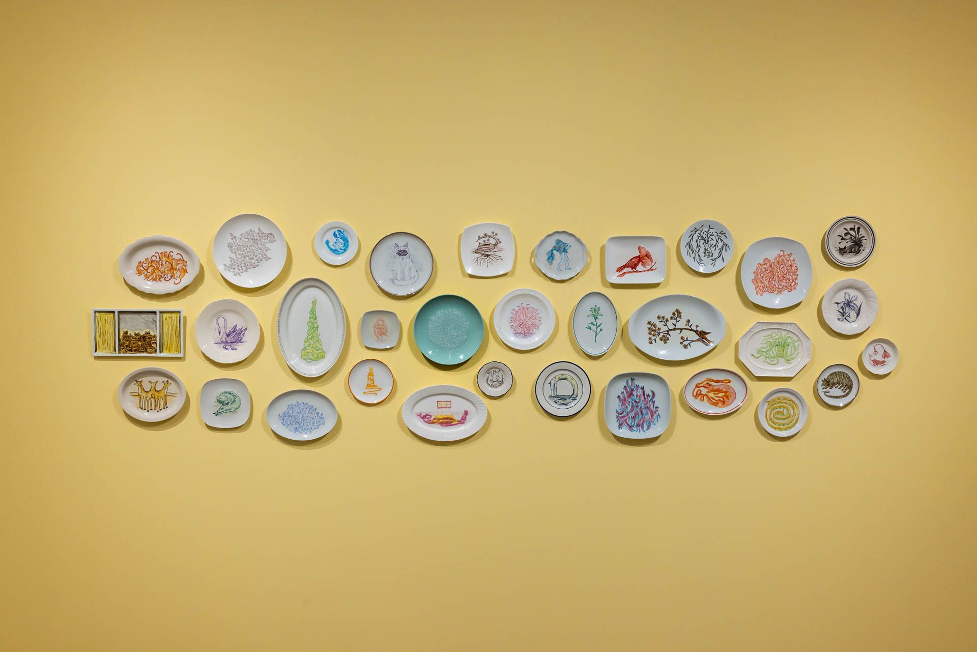 Cluster of painted plates and framed works arranged in a loose oval on a yellow wall. Installation view, Debra Broz: Strange World, Track 16.