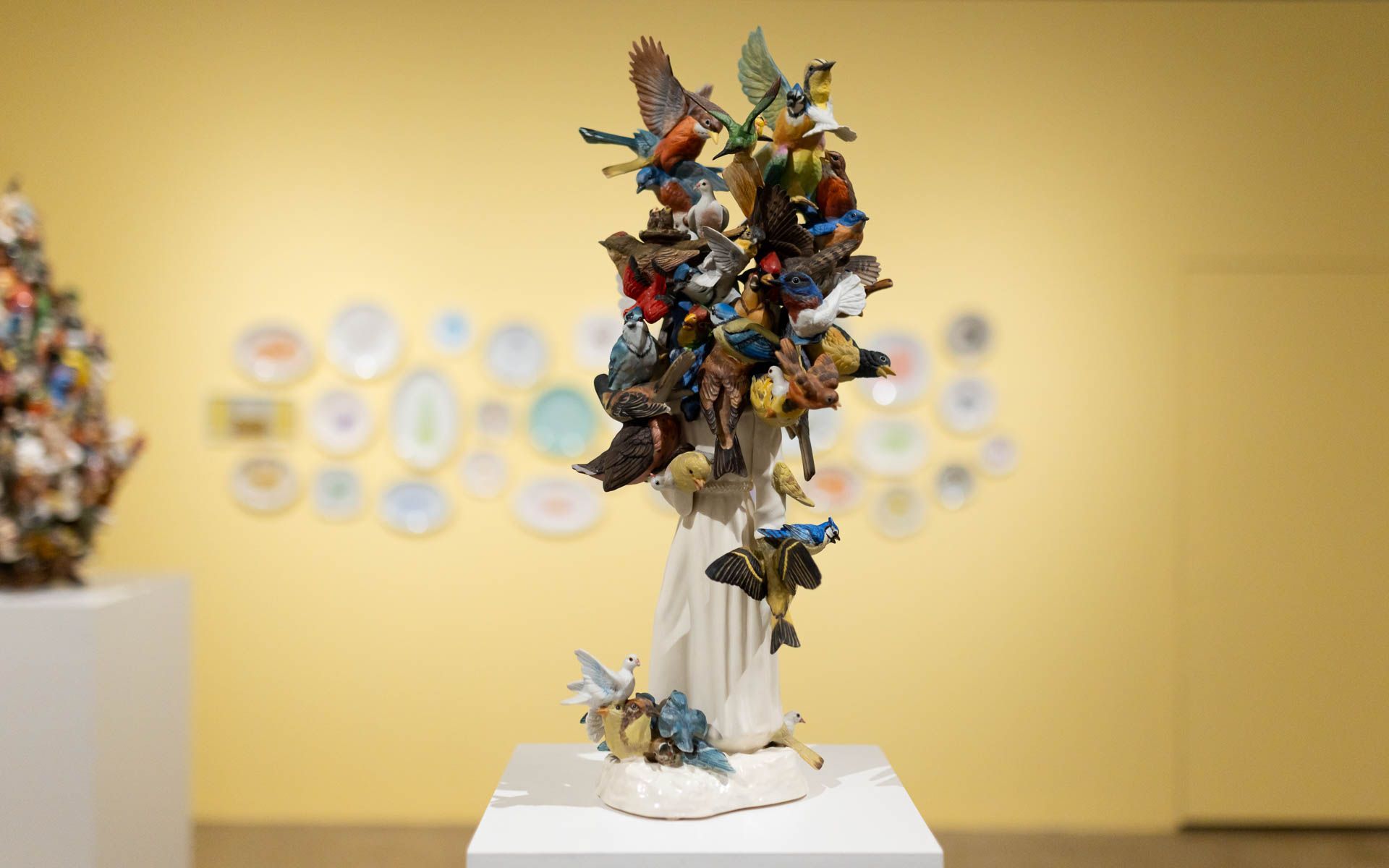 Ceramic sculpture of blue, red and brown birds perched on a white figure atop a pedestal, against a yellow wall of painted plates. Installation view, Debra Broz: Strange World, Track 16.