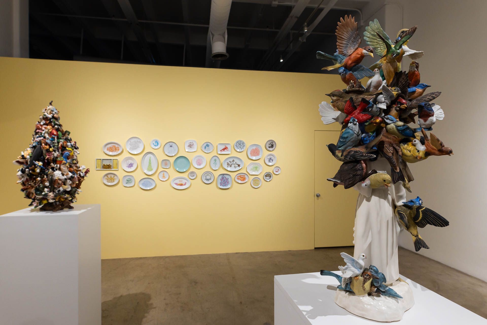 Ceramic tree of colorful animals on a pedestal at left, a yellow wall of painted plates and a sculpture of birds on a figure at right. Installation view, Debra Broz: Strange World, Track 16.