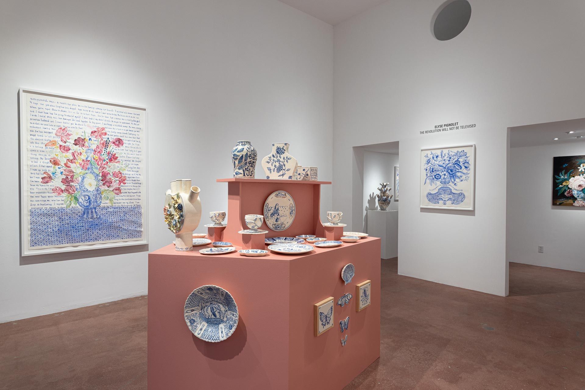 Art gallery with colorful mixed-media works on white walls and a pink display table in the center. Elyse Pignolet art exhibition at Track 16 East Hollywood gallery. Title, The Revolution Will Not Be Televised featuring ceramic works and drawings.