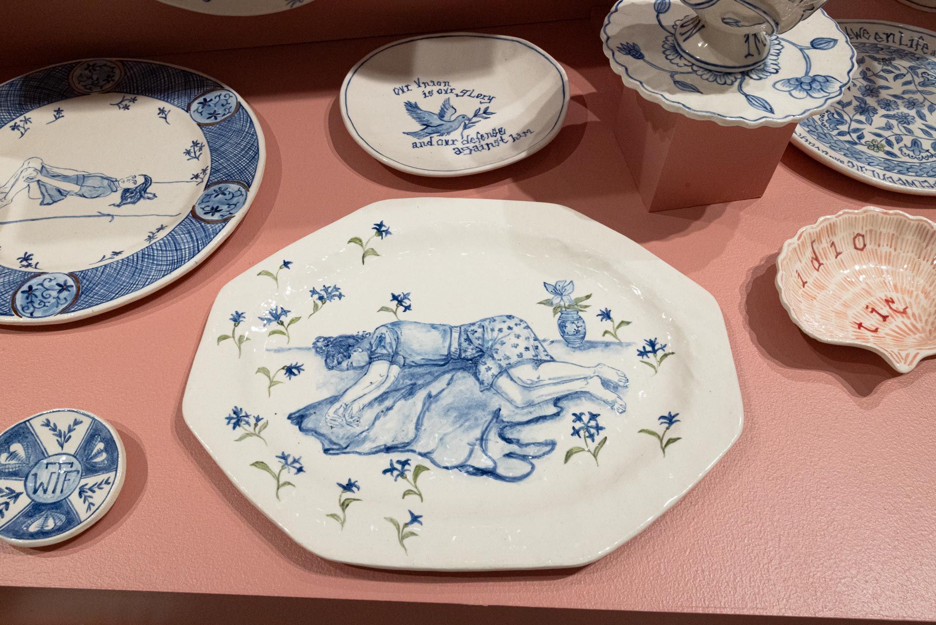 Blue-and-white ceramic plates and bowls arranged on a pink tablecloth. Elyse Pignolet art exhibition at Track 16 East Hollywood gallery. Title, The Revolution Will Not Be Televised featuring ceramic works and drawings.