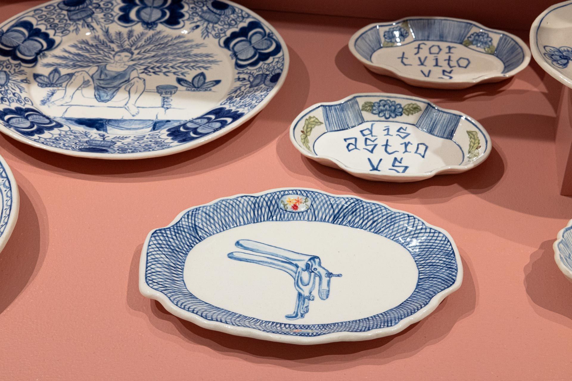 Blue-and-white decorative plates and small dishes displayed on a pink tabletop. Elyse Pignolet art exhibition at Track 16 East Hollywood gallery. Title, The Revolution Will Not Be Televised featuring ceramic works and drawings.
