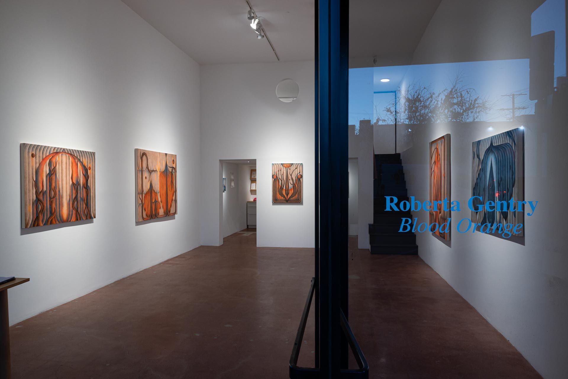 Modern art gallery corridor with framed paintings on white walls and blue text on the glass door. Roberta Gentry art exhibition at Track 16, East Hollywood in Los Angeles. Title , Blood Orange.