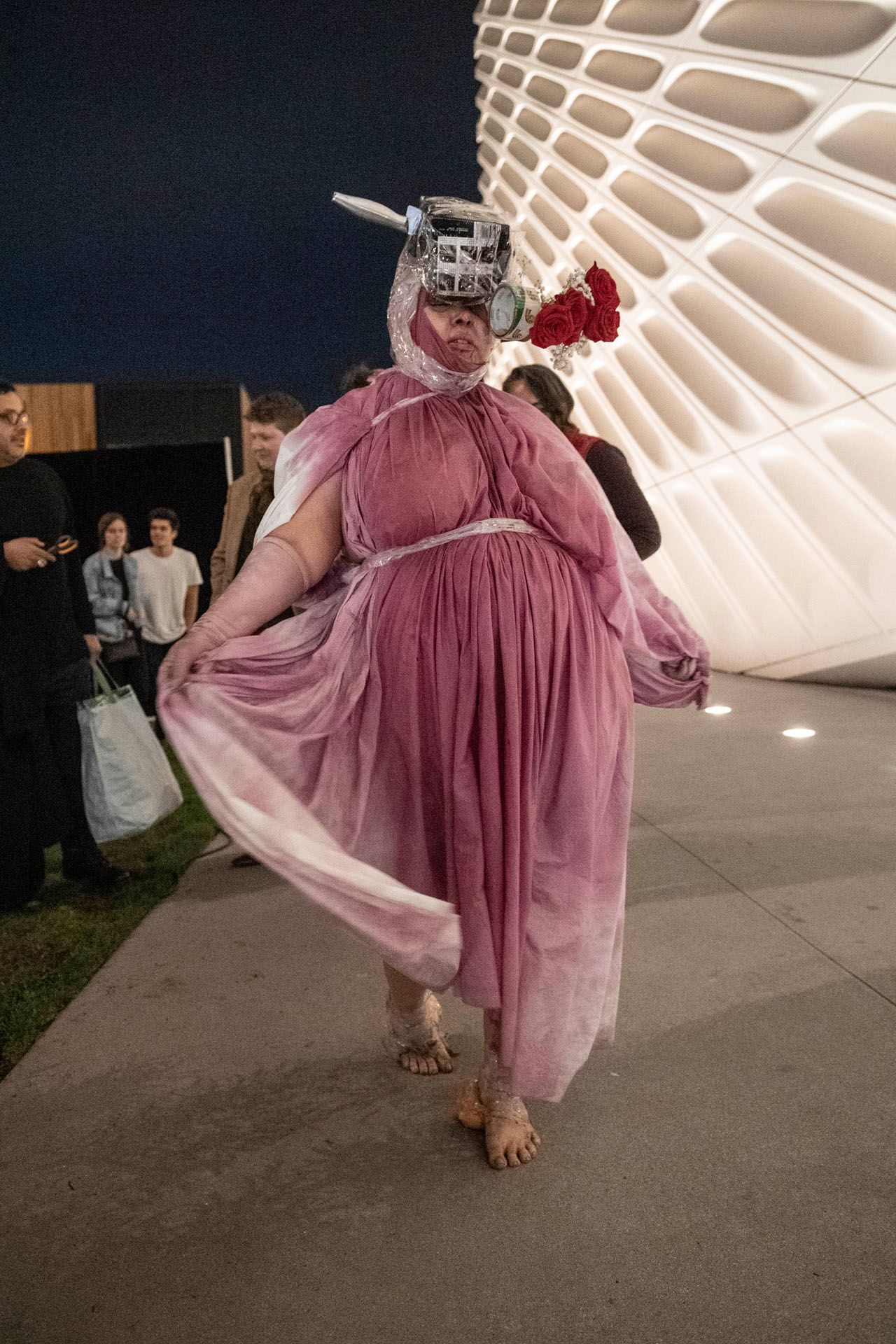 A woman in a pink dress and mask is standing on a sidewalk.