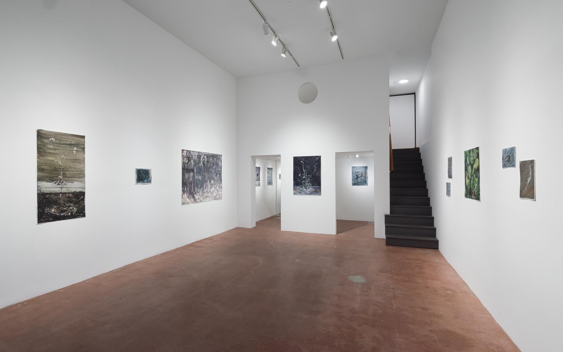 Tall dark gray panel and small paintings on a white wall, a doorway with a framed work beyond, and green works beside a staircase. Installation view, Molly Segal: As We Grind Our Teeth To Dust, Track 16.