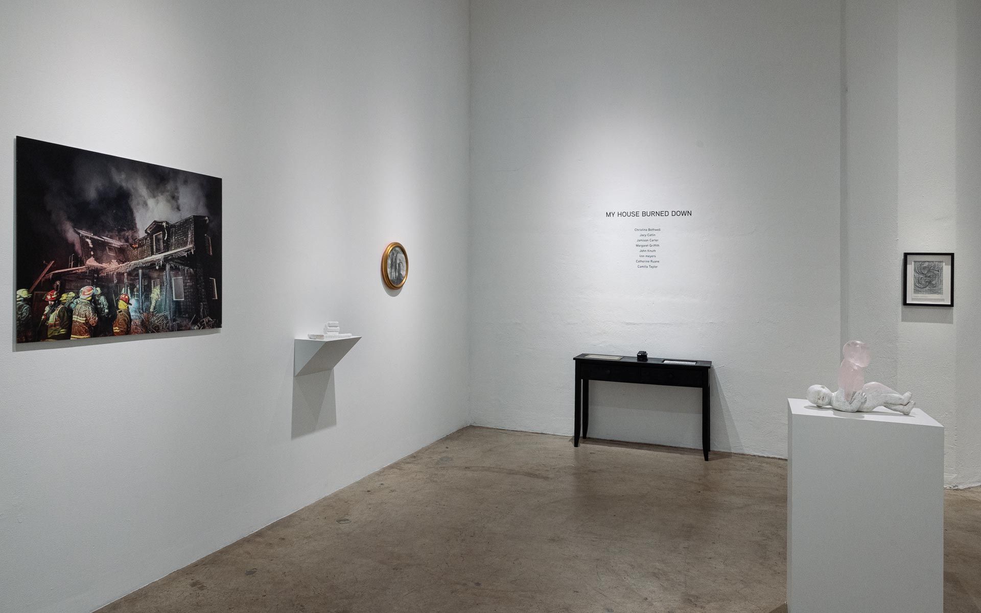 Large photograph of a burning building with firefighters at left, a small round framed work, a black console table and a white sculpture on a pedestal. Installation view, My House Burned Down, Track 16.