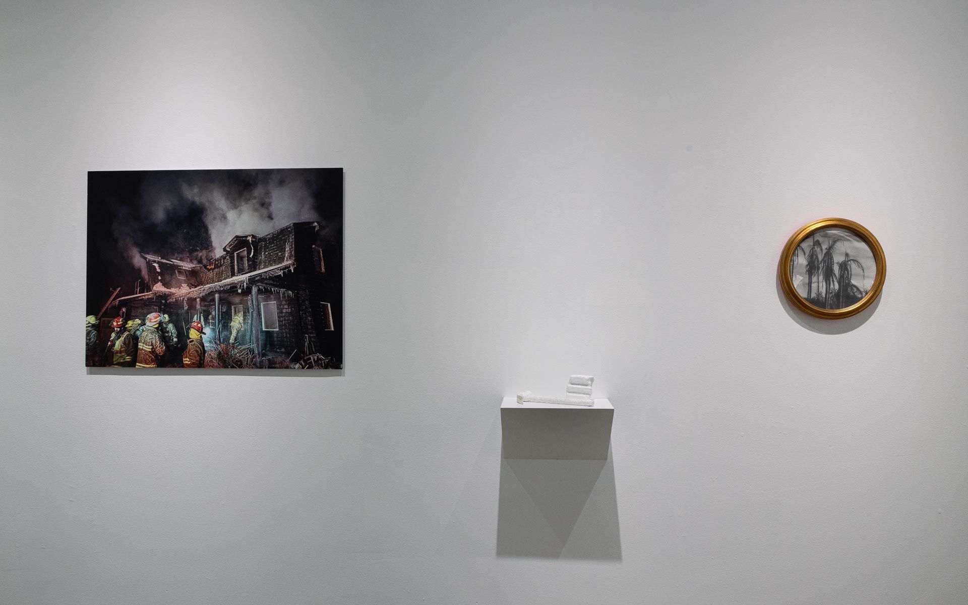 Large photograph of a burning building with firefighters, a white wall shelf and a round gold-framed work on a white wall. Installation view, My House Burned Down, Track 16.
