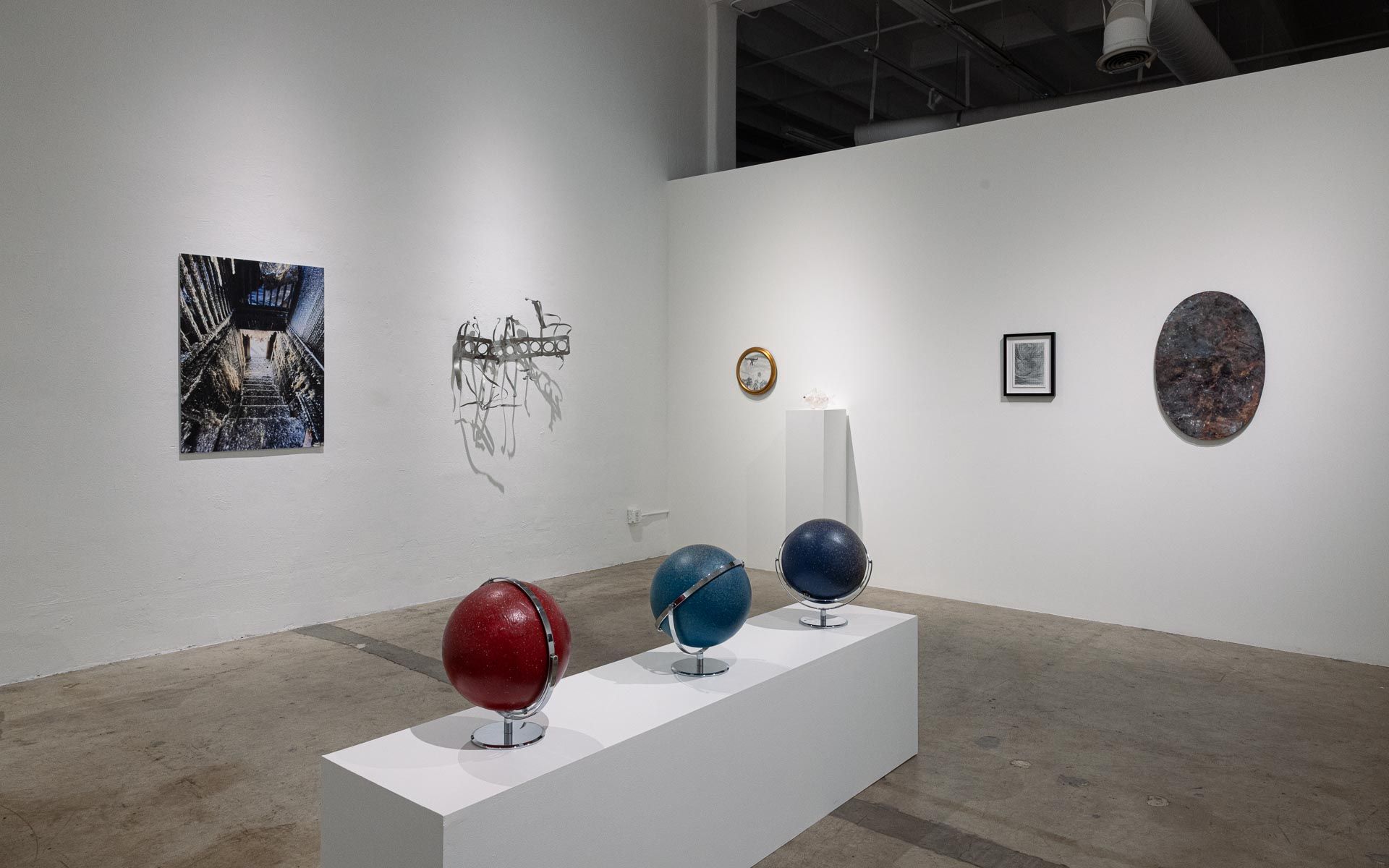 Red and blue globes on a long white plinth in the foreground, with framed works, a wire sculpture and a round framed piece on the white walls. Installation view, My House Burned Down, Track 16.