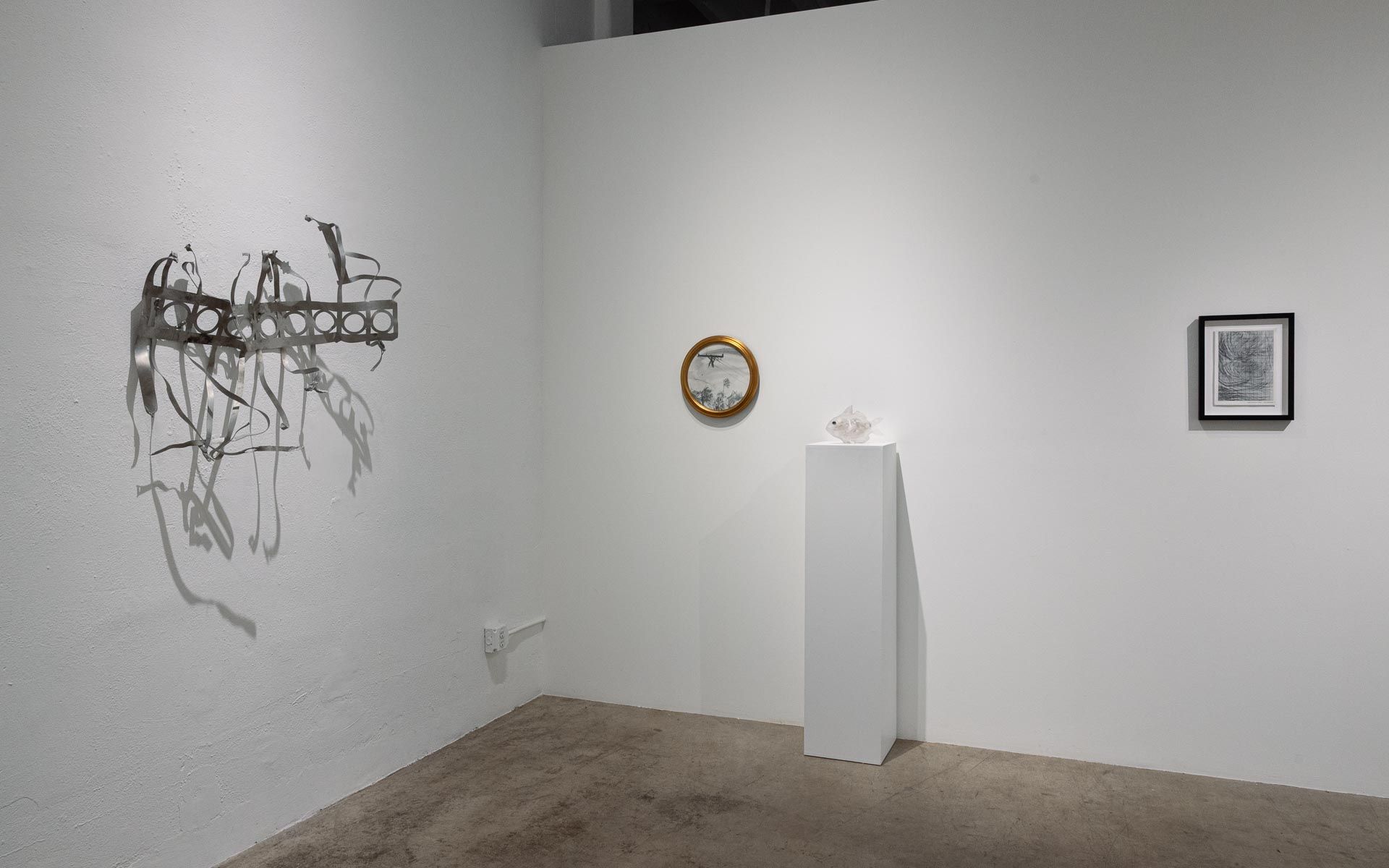 Gray wire and ribbon wall sculpture at left, a small round framed work above a white pedestal, and a framed print on the white wall. Installation view, My House Burned Down, Track 16.