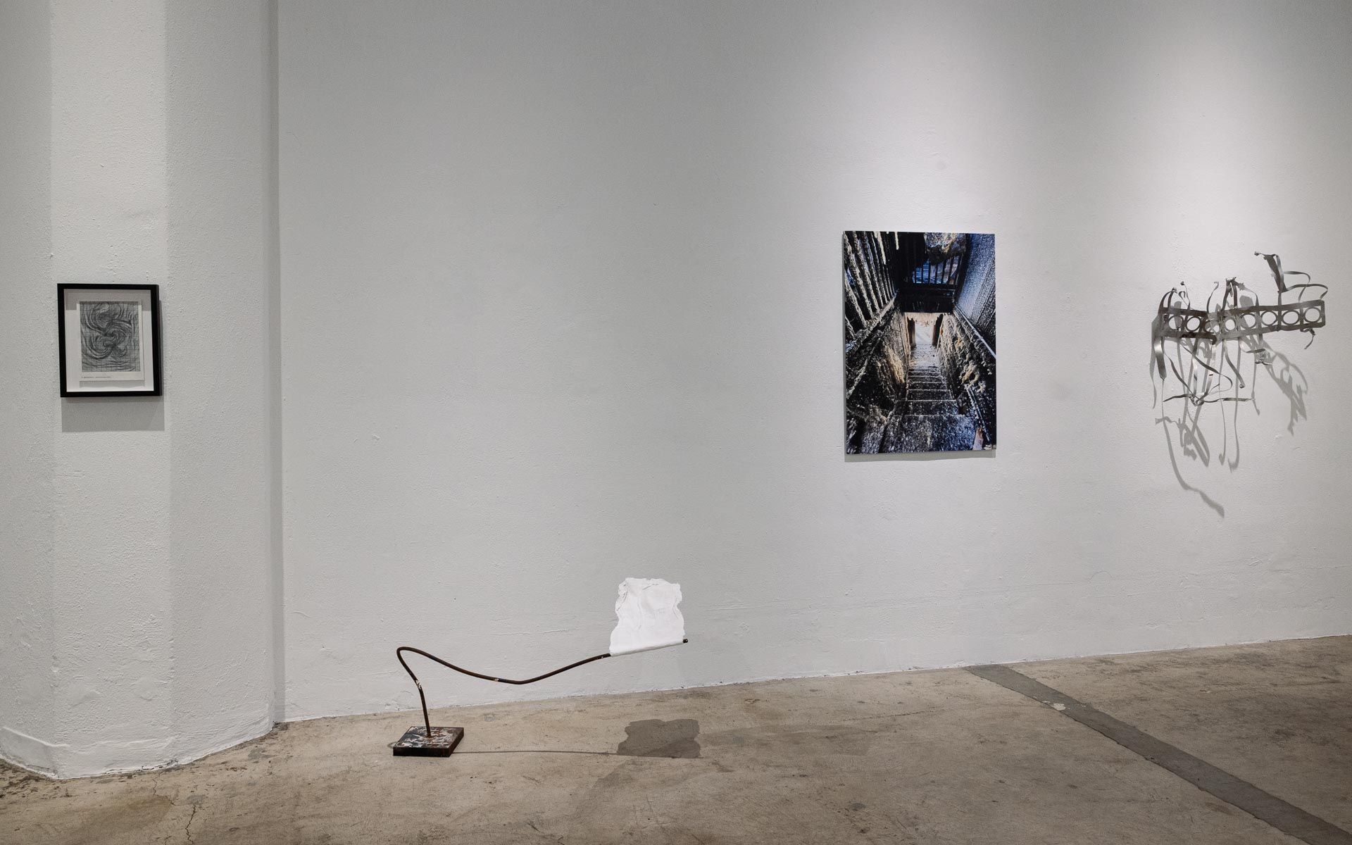 Framed print, a dark photograph and a gray wire sculpture on a white wall beside a column, with a cord trailing across the concrete floor. Installation view, My House Burned Down, Track 16.