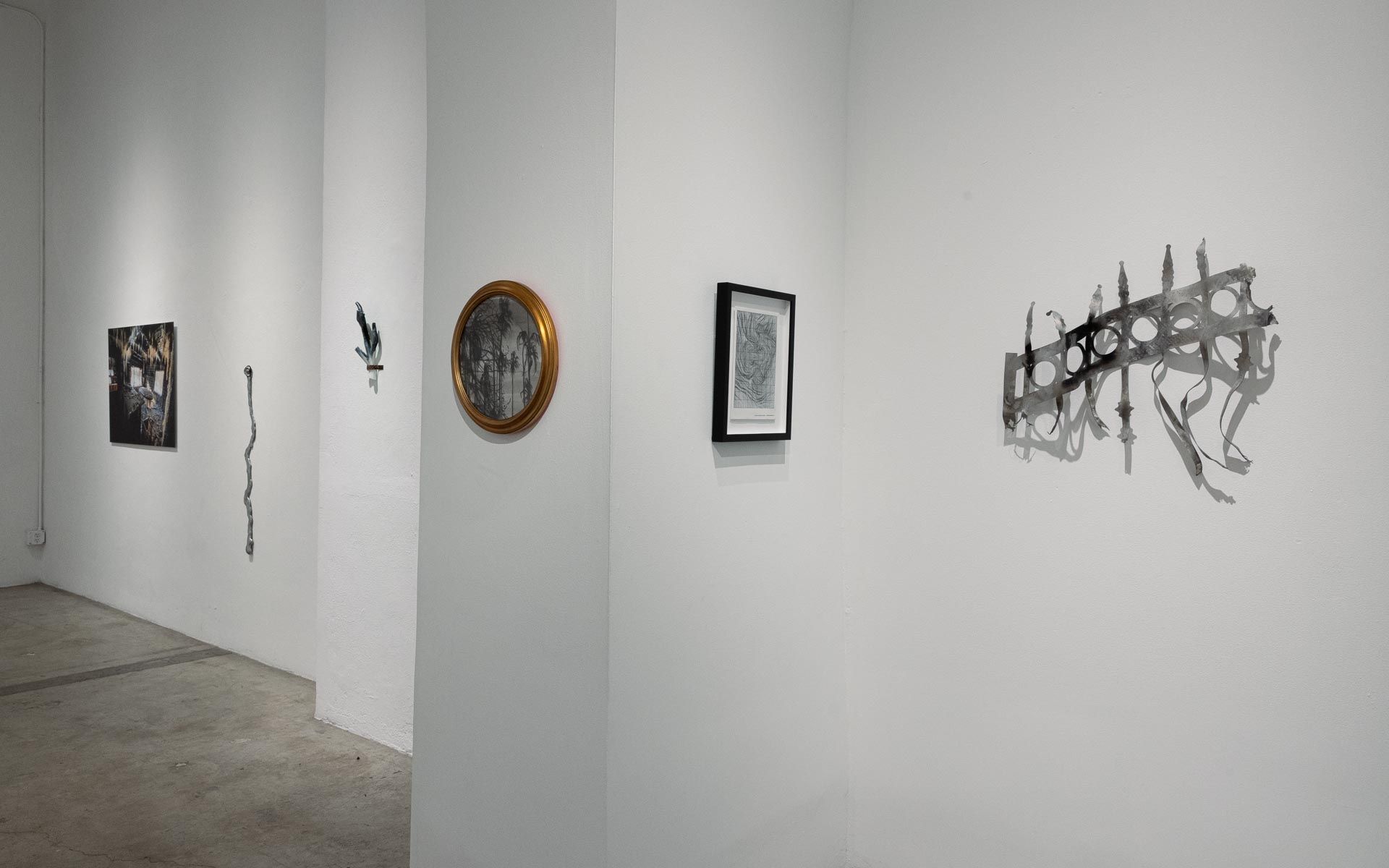 Round gold-framed work, a dark photograph, a framed print and a gray wire wall sculpture on white walls around a column. Installation view, My House Burned Down, Track 16.