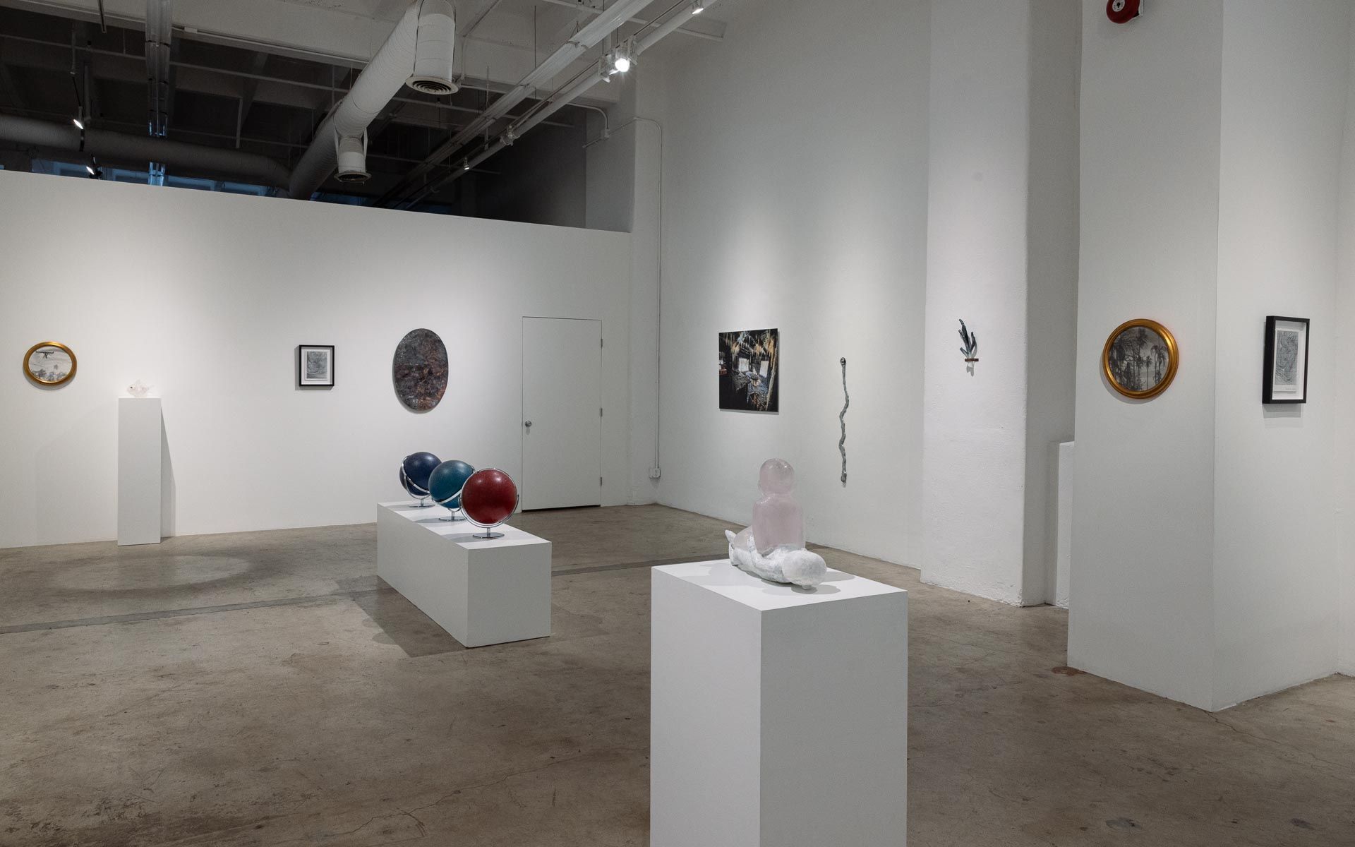 Wide view of a gallery with framed works on white walls, red and blue globes on a plinth and a white sculpture on a tall pedestal. Installation view, My House Burned Down, Track 16.