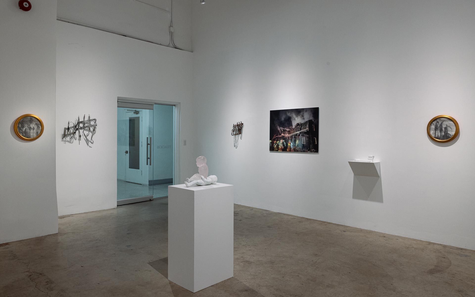 White sculpture on a pedestal, a photograph of a burning building and two round framed works on white walls around a glass door. Installation view, My House Burned Down, Track 16.