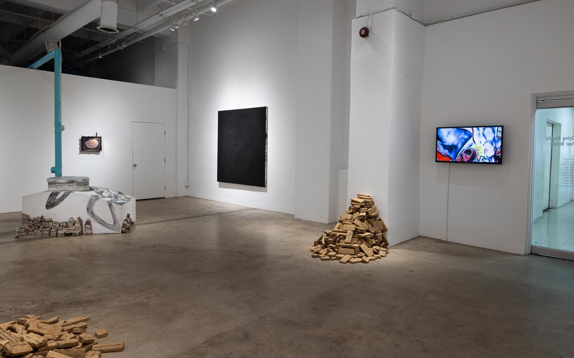 Modern art gallery with sculptures, wall-mounted artworks, and a video screen in a white exhibition room. Praise the Dying Beauty art exhibition at Track 16 Downtown Los Angeles gallery. Curated by Galia Linn featuring sculpture, paintings and drawings. 