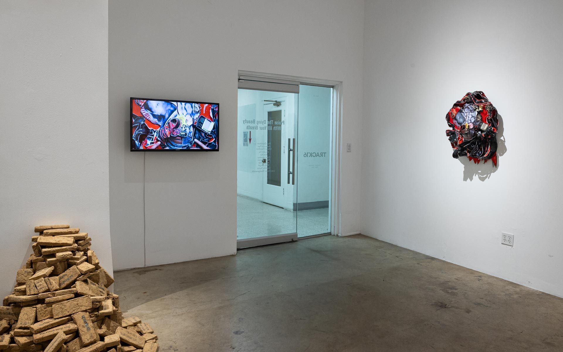 Art gallery room with a wall-mounted video, open doorway, wooden pile on floor, and a circular wall sculpture. Praise the Dying Beauty art exhibition at Track 16 Downtown Los Angeles gallery. Curated by Galia Linn featuring sculpture, paintings and drawings. 